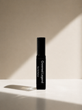 A single black cylindrical cosmetic bottle labeled 'Crowned Legend' and 'Eau de Parfum' stands upright on a white surface against a warm beige wall with dramatic diagonal sunlight and shadows.