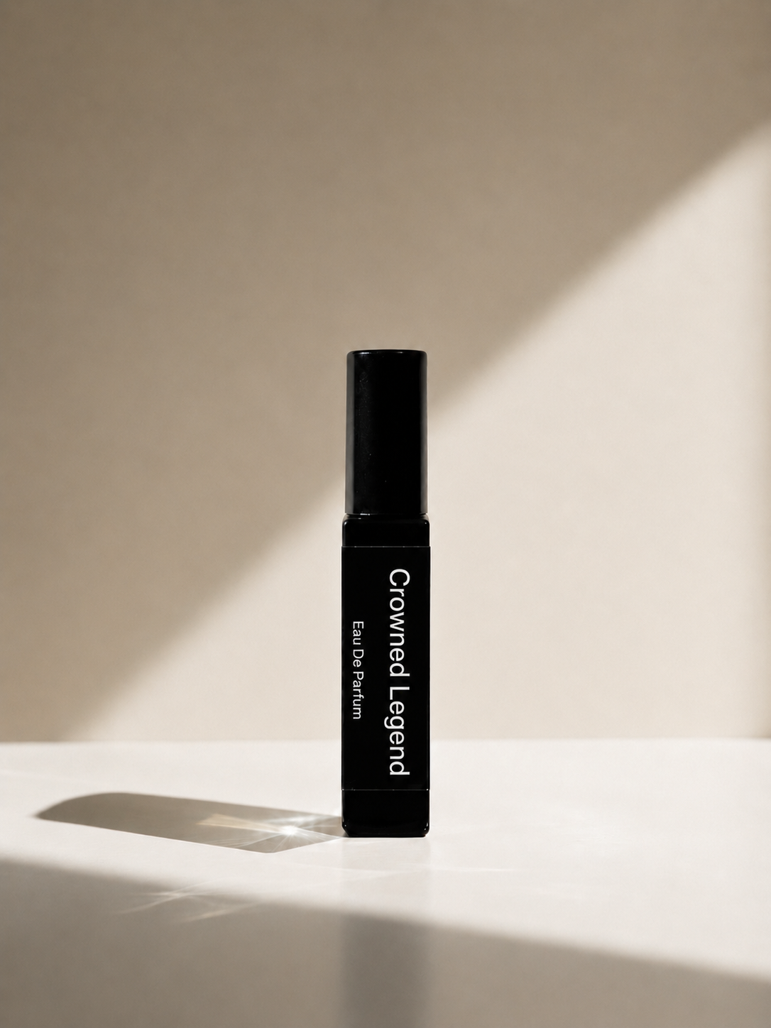 A single black cylindrical cosmetic bottle labeled 'Crowned Legend' and 'Eau de Parfum' stands upright on a white surface against a warm beige wall with dramatic diagonal sunlight and shadows.
