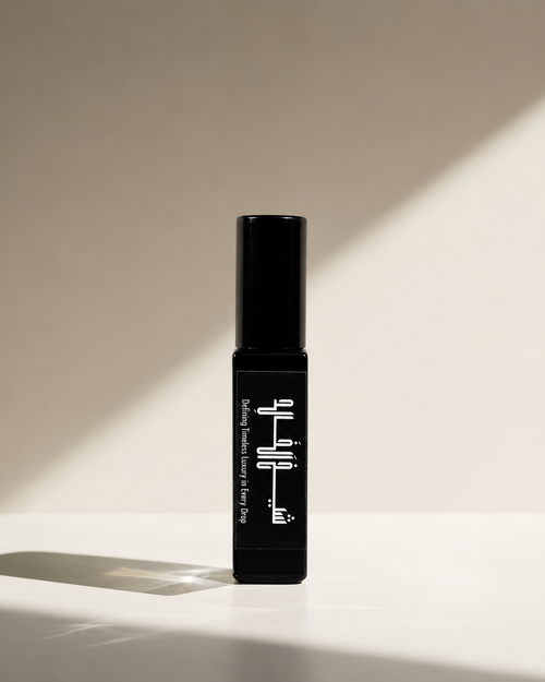 A black perfume bottle with white text stands upright on a white surface against a beige wall, lit by dramatic diagonal sunlight that casts strong shadows.