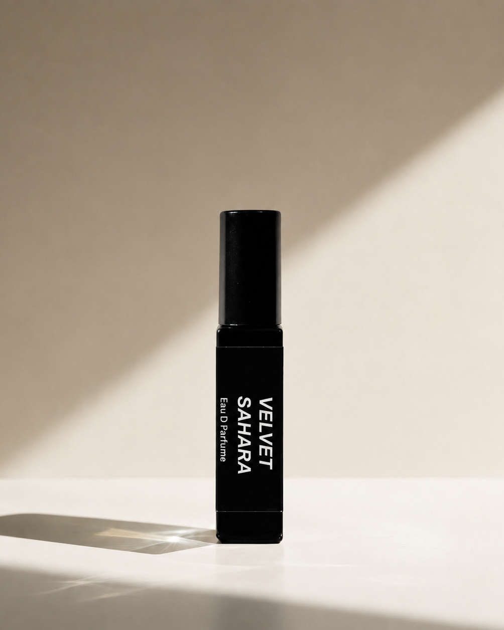 A black cylindrical perfume bottle labeled 'VELVET SAHARA Eau De Parfum' stands upright on a white surface with dramatic diagonal sunlight casting shadows on a beige background.