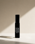 A black cylindrical perfume bottle labeled 'VELVET SAHARA Eau De Parfum' stands upright on a white surface with dramatic diagonal sunlight casting shadows on a beige background.